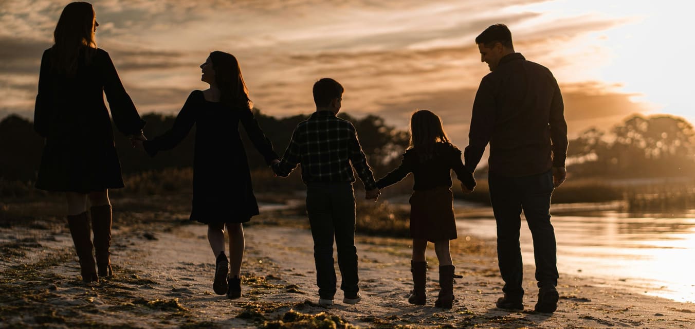 Una famiglia per mano, di spalle, su una spiaggia al tramonto